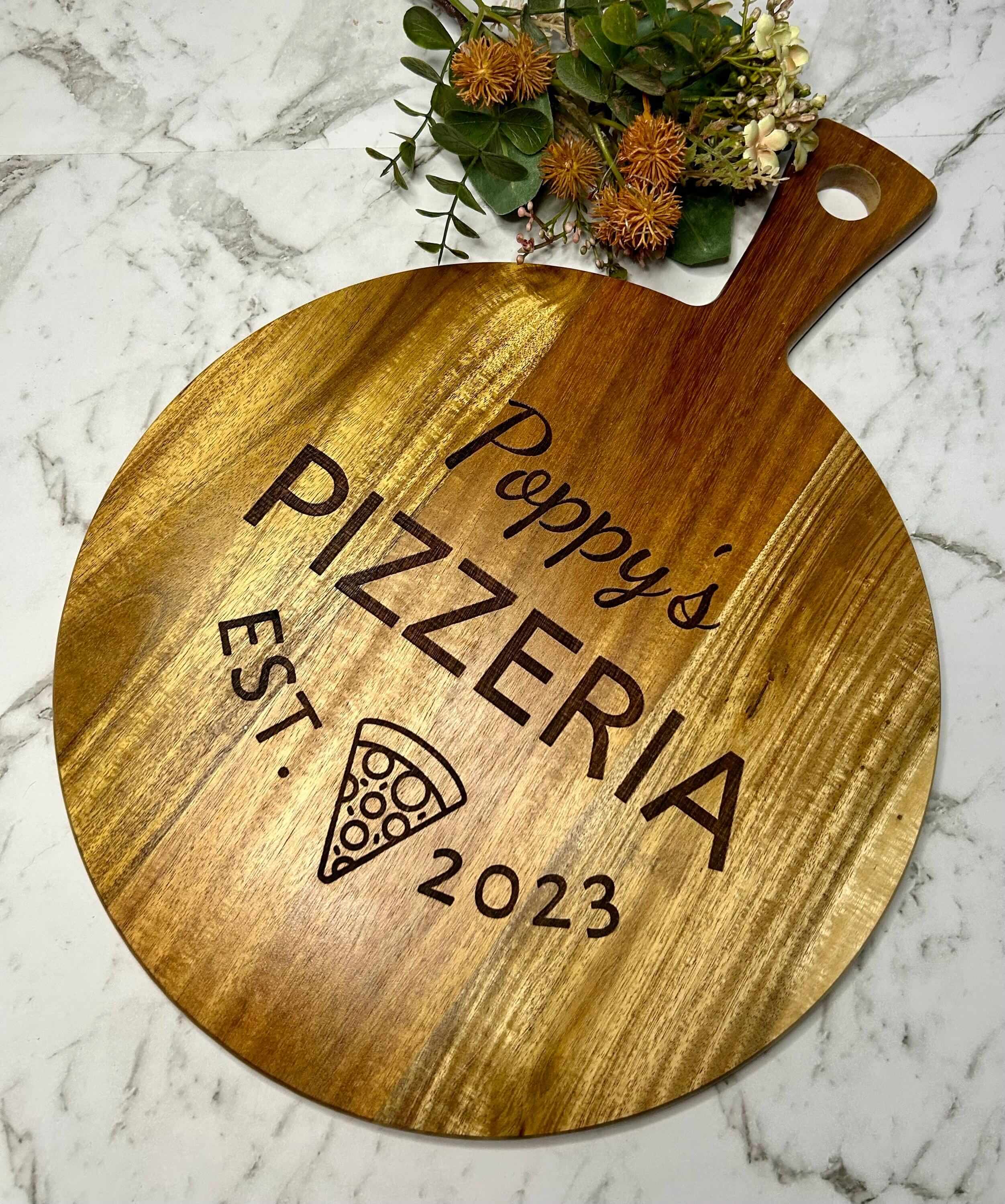 Acacia wood pizza board showing engraving of a name followed by PIZZERIA, est and a date. emoji of pizza slice below pizzeria